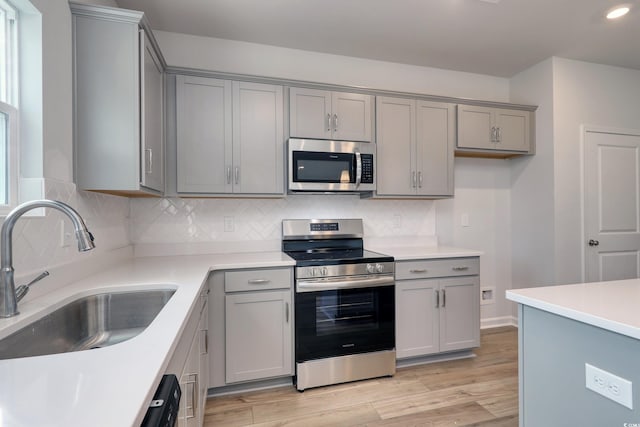 kitchen with gray cabinetry, decorative backsplash, a sink, light wood-style flooring, and appliances with stainless steel finishes