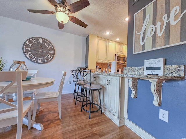 kitchen with light wood-type flooring, a textured ceiling, a breakfast bar area, light stone countertops, and ceiling fan