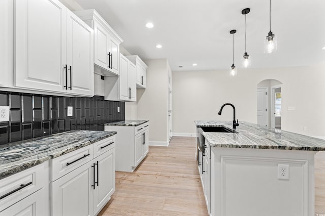 kitchen featuring light wood finished floors, a center island with sink, arched walkways, decorative backsplash, and light stone countertops