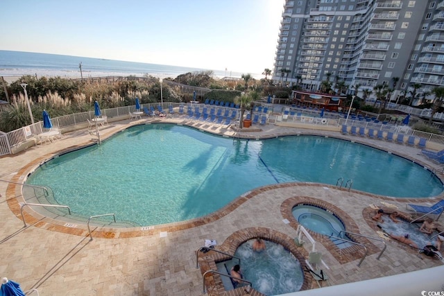 pool with a view of the beach, a community hot tub, a water view, a patio area, and fence