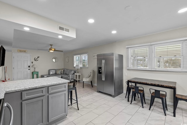 kitchen with stainless steel fridge with ice dispenser, a breakfast bar, gray cabinets, ceiling fan, and light stone counters