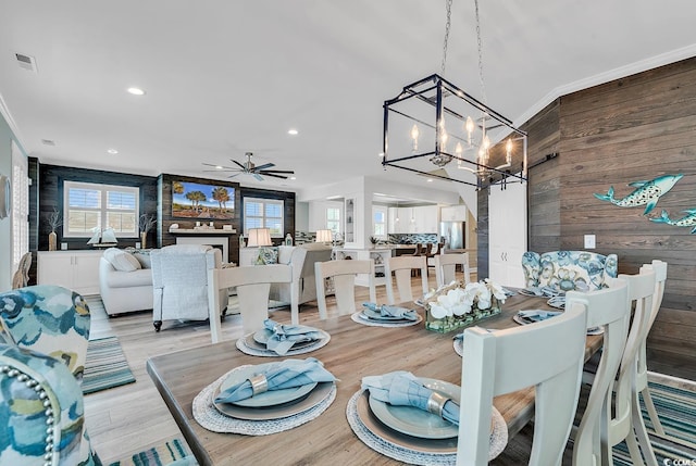 dining area with ceiling fan with notable chandelier, crown molding, wooden walls, and light wood-type flooring