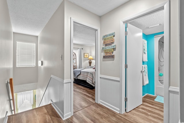 hallway featuring a wainscoted wall, light wood finished floors, a textured ceiling, and an upstairs landing