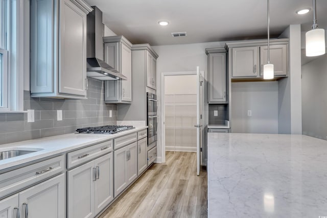 kitchen with light stone counters, gray cabinetry, stainless steel appliances, wall chimney range hood, and pendant lighting