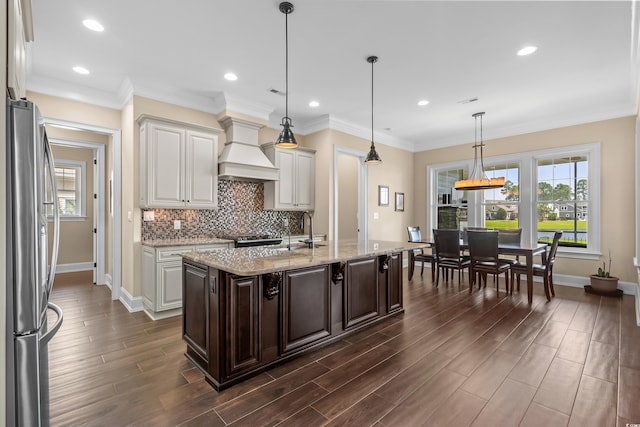 kitchen featuring a kitchen island with sink, stainless steel refrigerator, custom range hood, sink, and decorative light fixtures