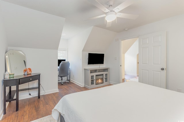 bedroom with baseboards, lofted ceiling, a warm lit fireplace, wood finished floors, and a ceiling fan