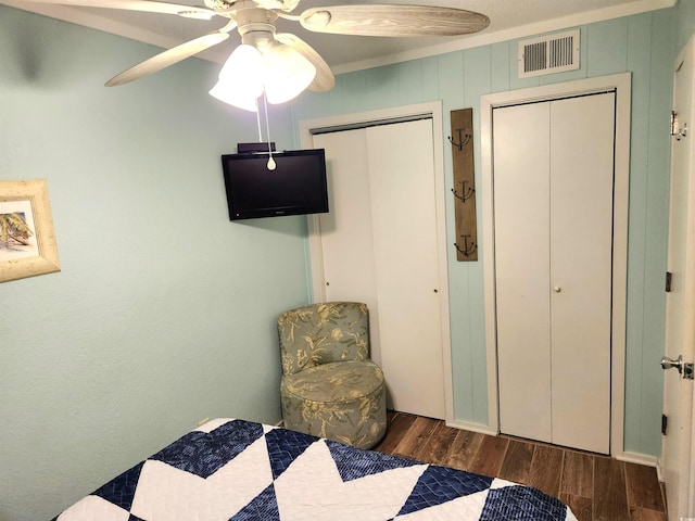 bedroom with ceiling fan, wood finished floors, visible vents, ornamental molding, and two closets