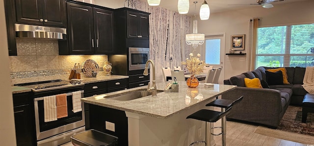 kitchen with pendant lighting, ventilation hood, an island with sink, sink, and stainless steel appliances