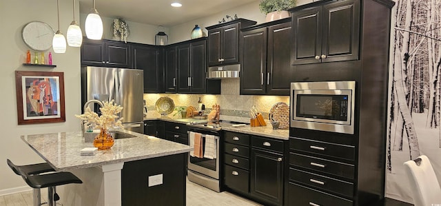 kitchen featuring backsplash, appliances with stainless steel finishes, light stone counters, a breakfast bar area, and a center island with sink