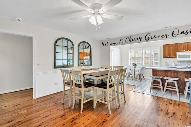 dining room with light wood-style floors, a textured ceiling, a ceiling fan, and wainscoting