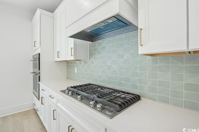 kitchen with white cabinetry, stainless steel appliances, custom range hood, light hardwood / wood-style flooring, and backsplash