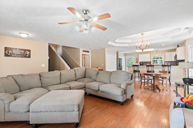 living room with light wood-type flooring, ceiling fan with notable chandelier, a raised ceiling, and a textured ceiling