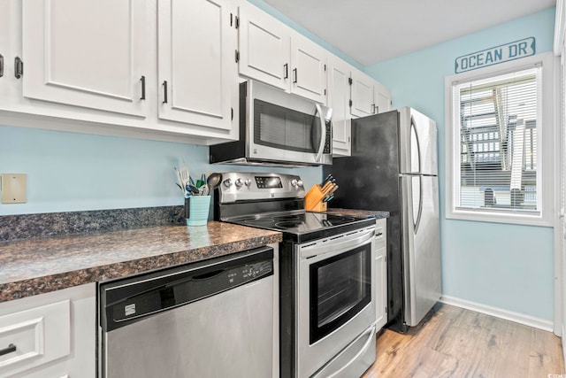 kitchen with dark countertops, light wood-style flooring, appliances with stainless steel finishes, white cabinets, and baseboards