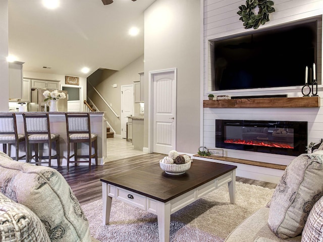 living area with light wood-type flooring, a glass covered fireplace, lofted ceiling, and stairs