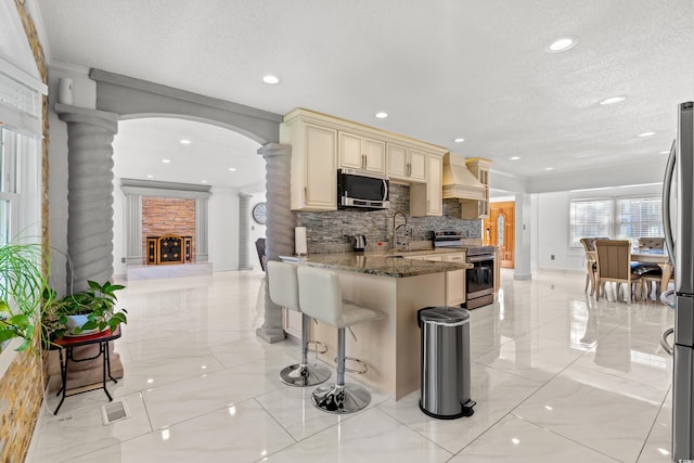 kitchen with custom exhaust hood, stone counters, decorative backsplash, appliances with stainless steel finishes, and cream cabinetry