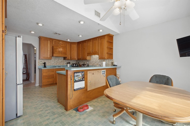 kitchen with a peninsula, light countertops, freestanding refrigerator, brown cabinets, and tasteful backsplash