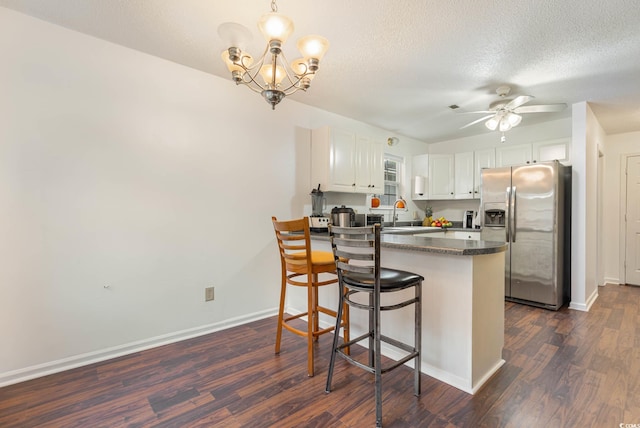 kitchen with dark wood-type flooring, ceiling fan with notable chandelier, stainless steel refrigerator with ice dispenser, white cabinetry, and a kitchen breakfast bar