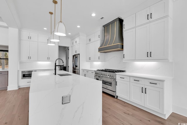 kitchen featuring premium range hood, hanging light fixtures, sink, light wood-type flooring, and appliances with stainless steel finishes