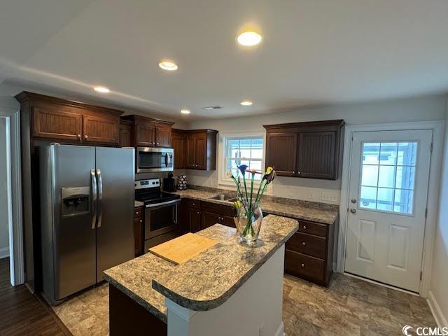 kitchen with appliances with stainless steel finishes, light stone counters, dark brown cabinets, sink, and a kitchen island