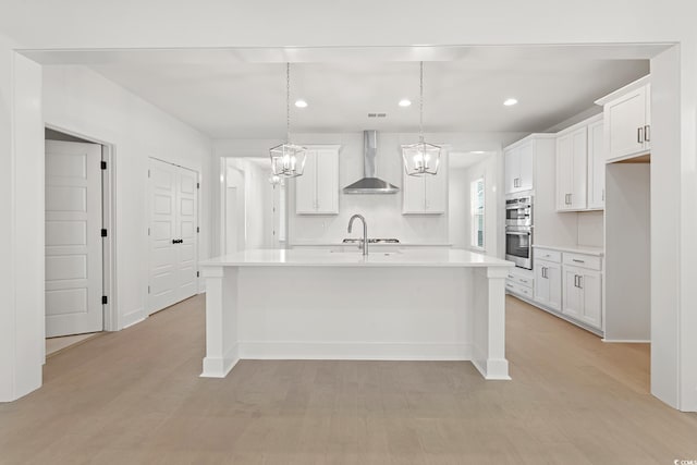 kitchen with wall chimney exhaust hood, white cabinets, an island with sink, and double oven