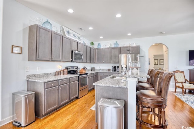 kitchen with light stone counters, a center island with sink, stainless steel appliances, a breakfast bar area, and light hardwood / wood-style floors