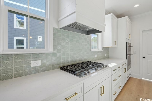 kitchen featuring custom exhaust hood, appliances with stainless steel finishes, light wood-type flooring, ornamental molding, and white cabinets