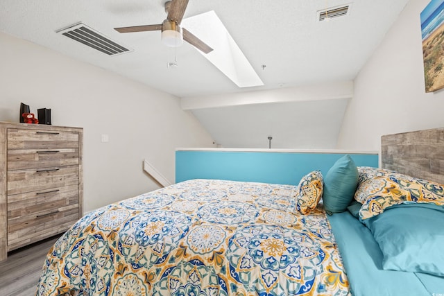 bedroom with a textured ceiling, vaulted ceiling with skylight, ceiling fan, and light hardwood / wood-style flooring