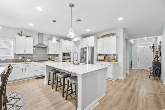 kitchen featuring white cabinets, an island with sink, wall chimney range hood, pendant lighting, and appliances with stainless steel finishes