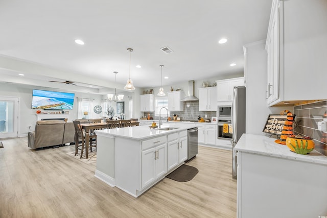 kitchen featuring stainless steel appliances, white cabinetry, wall chimney range hood, sink, and an island with sink