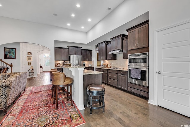 kitchen featuring stainless steel appliances, arched walkways, dark brown cabinetry, a breakfast bar area, and a towering ceiling