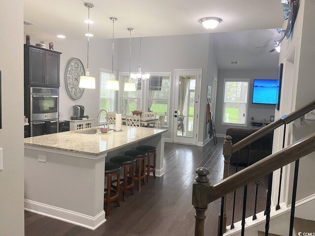 kitchen with sink, a kitchen island with sink, light stone countertops, decorative light fixtures, and stainless steel double oven