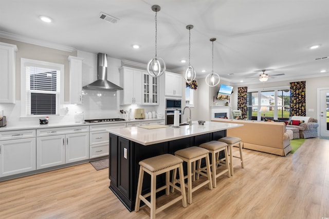 kitchen with wall chimney exhaust hood, white cabinetry, a center island with sink, and light hardwood / wood-style floors