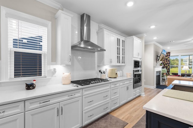 kitchen featuring light wood-type flooring, white cabinets, wall chimney range hood, appliances with stainless steel finishes, and ornamental molding