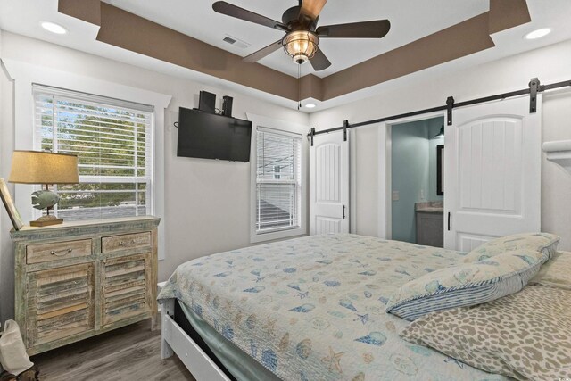 bedroom with ensuite bathroom, a raised ceiling, ceiling fan, a barn door, and wood-type flooring