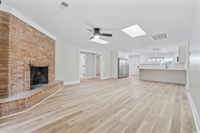 unfurnished living room with light wood-type flooring, a skylight, a brick fireplace, and ceiling fan