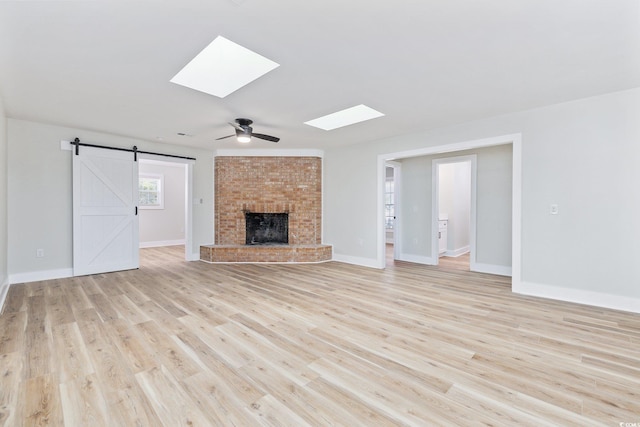 unfurnished living room with a skylight, ceiling fan, a brick fireplace, a barn door, and light wood-type flooring