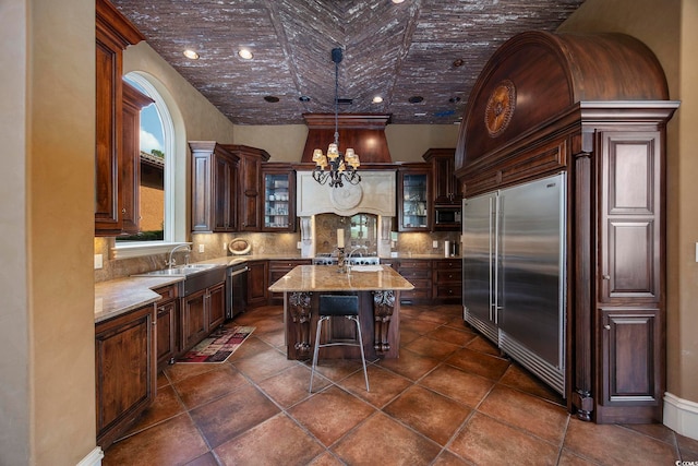 kitchen featuring a notable chandelier, a kitchen island with sink, backsplash, glass insert cabinets, and built in appliances