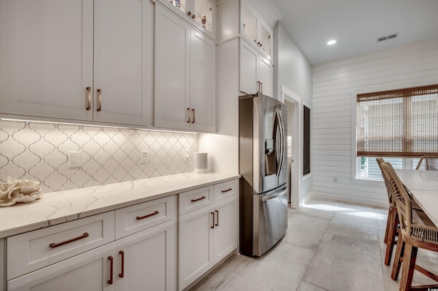 kitchen featuring stainless steel fridge, light stone counters, wooden walls, backsplash, and white cabinetry