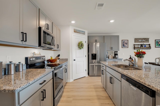 kitchen with gray cabinetry, stainless steel appliances, sink, light stone counters, and light hardwood / wood-style flooring