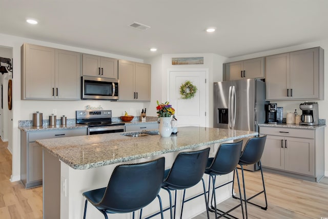 kitchen with light wood-type flooring, appliances with stainless steel finishes, a kitchen breakfast bar, an island with sink, and light stone counters