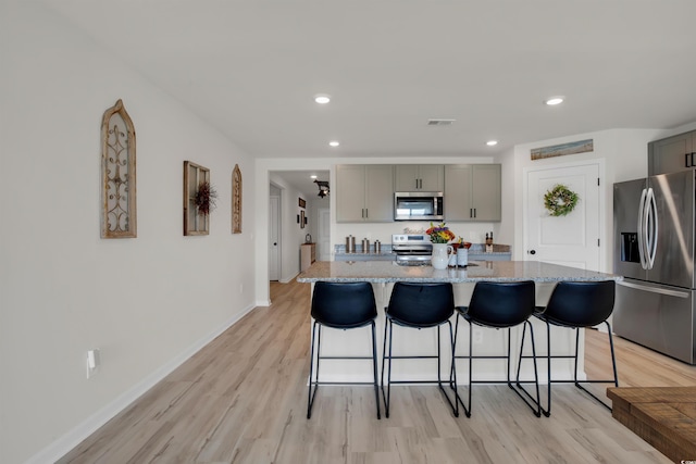 kitchen featuring a center island with sink, a breakfast bar area, stainless steel appliances, gray cabinetry, and light stone counters