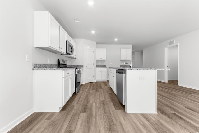 kitchen with an island with sink, appliances with stainless steel finishes, white cabinetry, light hardwood / wood-style floors, and light stone counters