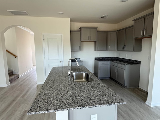 kitchen with a kitchen island with sink, sink, dark stone countertops, and light wood-type flooring