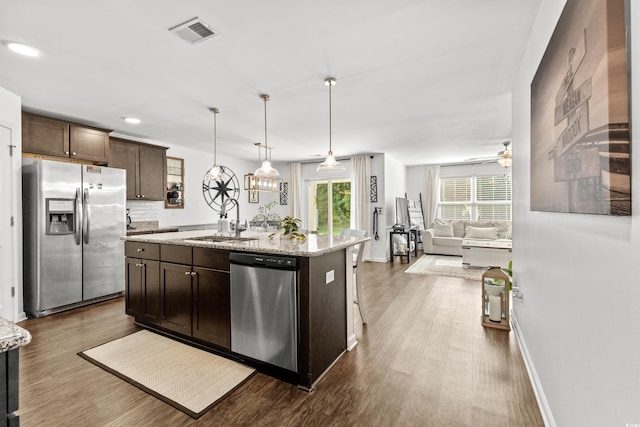 kitchen featuring stainless steel appliances, a center island with sink, dark brown cabinetry, light stone counters, and dark hardwood / wood-style flooring