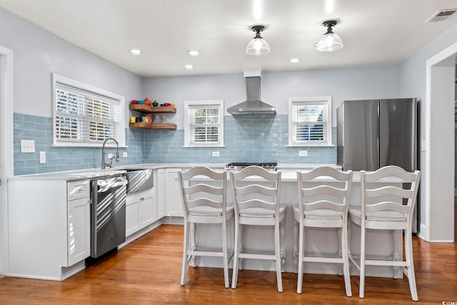 kitchen featuring wall chimney range hood, light hardwood / wood-style floors, stainless steel appliances, decorative light fixtures, and white cabinets