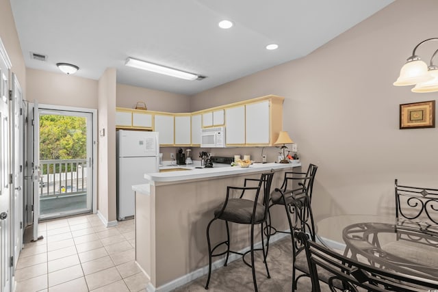 kitchen featuring kitchen peninsula, light tile patterned flooring, a kitchen bar, white cabinetry, and white appliances