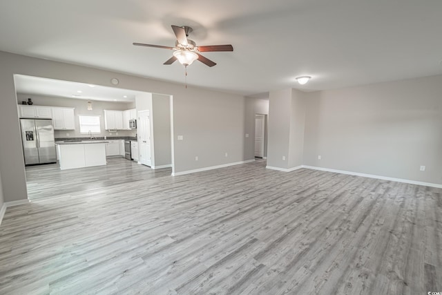 unfurnished living room with ceiling fan, a sink, light wood-style flooring, and baseboards