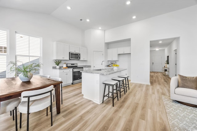 kitchen with a center island with sink, appliances with stainless steel finishes, white cabinetry, light wood-type flooring, and light stone counters