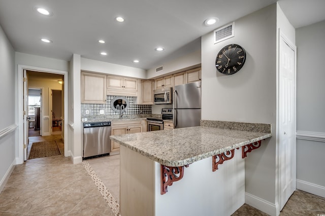 kitchen featuring kitchen peninsula, backsplash, appliances with stainless steel finishes, light stone countertops, and light brown cabinetry