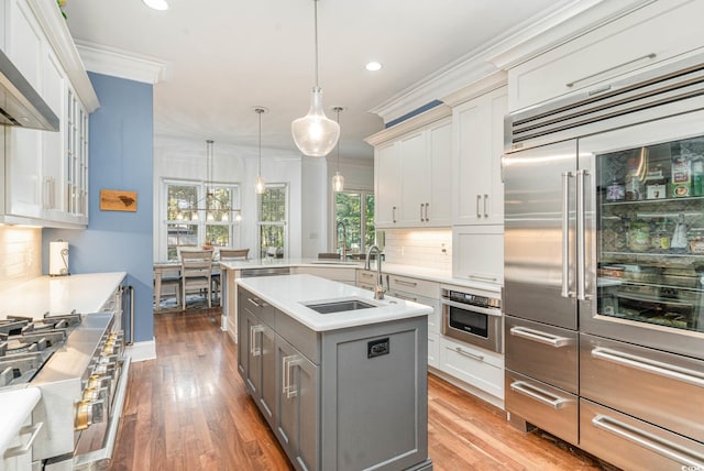 kitchen with high end appliances, an island with sink, wall chimney exhaust hood, light countertops, and crown molding